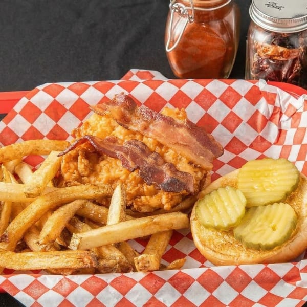 Crispy chicken sandwich with bacon and pickles, served with fries in a red checkered basket.