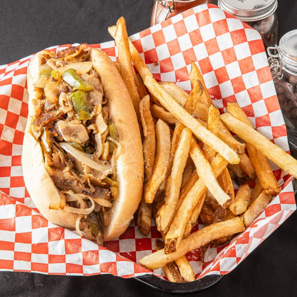 Cheesesteak sandwich with onions and peppers, served with a side of golden french fries in a red checkered basket.