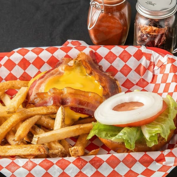 A bacon cheeseburger with lettuce, tomato, and onion, served with fries in a red and white checkered basket.